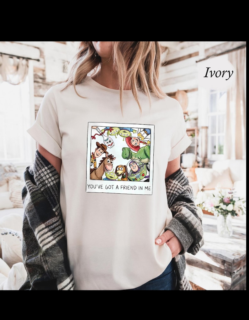 Op de afbeelding: Ivoorkleurig T-shirt met een ingelijste cartoonafbeelding van Toy Story-personages, waaronder Woody en Buzz Lightyear. De tekst "YOU'VE GOT A FRIEND IN ME" staat onder de afbeelding. Het T-shirt is licht cr&egrave;mekleurig.