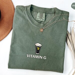 May include: A moss green t-shirt with a crew neck and short sleeves. The shirt features a graphic of a pint of beer with the text "VITAMIN G" in white. A gold necklace with a pendant is draped across the shirt.