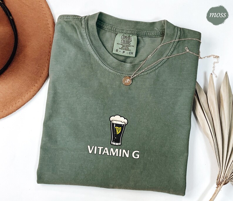 May include: A moss green t-shirt with a crew neck and short sleeves. The shirt features a graphic of a pint of beer with the text "VITAMIN G" in white. A gold necklace with a pendant is draped across the shirt.