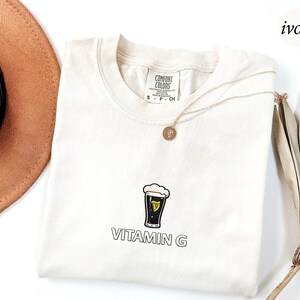 May include: Ivory-colored t-shirt with a graphic of a pint of stout beer and the text "VITAMIN G". The shirt has a crew neck and short sleeves. A gold necklace with a pendant is draped on the shirt.