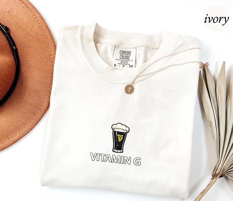 May include: Ivory-colored t-shirt with a graphic of a pint of stout beer and the text "VITAMIN G". The shirt has a crew neck and short sleeves. A gold necklace with a pendant is draped on the shirt.