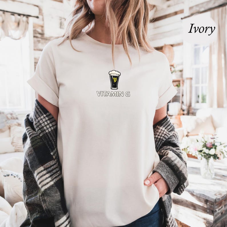 May include: Ivory-colored t-shirt with a graphic of a pint of stout beer and the words "VITAMIN G" printed below. The shirt is a crew neck style with rolled-up sleeves. The model is wearing a plaid shirt over the t-shirt.