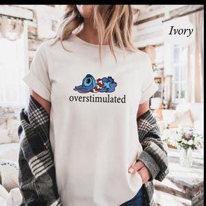 以下が含まれることがあります： アイボリー色のTシャツ。青、赤、白の漫画のキャラクターと、下に「overstimulated」の文字がプリントされています。Tシャツは半袖でゆったりとしたフィット感です。背景はぼやけています。