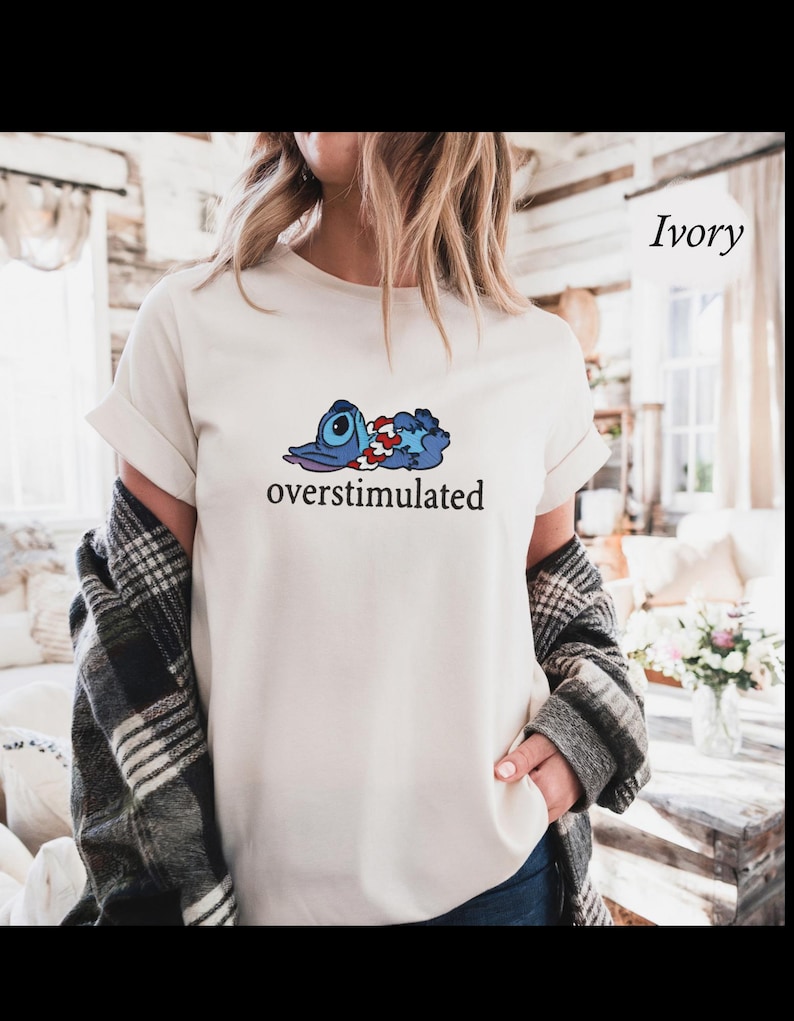 以下が含まれることがあります： アイボリー色のTシャツ。青、赤、白の漫画のキャラクターと、下に「overstimulated」の文字がプリントされています。Tシャツは半袖でゆったりとしたフィット感です。背景はぼやけています。