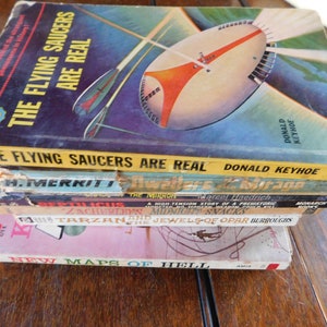 May include: A stack of vintage paperback books with colorful covers. The top book is titled "The Flying Saucers Are Real" by Donald Keyhoe. The other books include titles like "Reptilicus", "The Mirror", "Tarzan and the Jewels of Opar", and "New Maps of Hell".