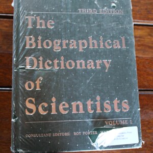Pode incluir: Um livro verde de capa dura intitulado "The Biographical Dictionary of Scientists" com as palavras "Third Edition" e "Volume I" na capa.