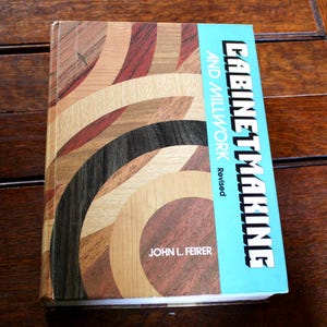 Può includere: Un libro intitolato "Cabinetmaking and Millwork Revised" di John L. Feirer. La copertina presenta un disegno di venature del legno curve in vari colori. Il dorso è azzurro con testo bianco.