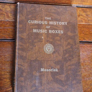 Puede incluir: Un libro antiguo con una cubierta marrón, titulado "The Curious History of Music Boxes". El título está en letras en relieve. El nombre del autor, Mosoriak, está impreso en la parte inferior. Un emblema decorativo está centrado en la cubierta.