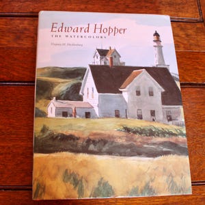 Puede incluir: Portada de libro con el título "Edward Hopper The Watercolors" y una pintura a la acuarela de una escena costera. La obra representa un faro blanco y edificios sobre una colina cubierta de hierba bajo un cielo claro. También se ve el nombre de la autora, Virginia M. Mecklenburg.