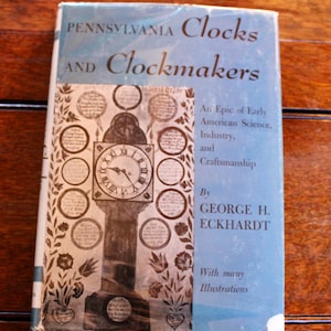 Puede incluir: Un libro antiguo titulado "Pennsylvania Clocks and Clockmakers" con una cubierta azul claro. La portada presenta una ilustración detallada de un reloj y elementos decorativos. El libro es de George H. Eckhardt.