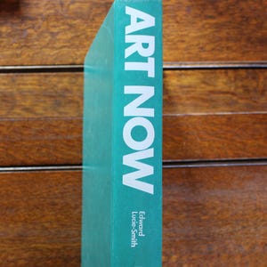 Może przedstawiać: Turkusowy grzbiet książki z tytułem "ART NOW" w dużych białych literach. Imię i nazwisko autora, Edward Lucie-Smith, wydrukowane poniżej. Grzbiet książki na tle drewnianego tła.