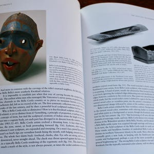 以下が含まれることがあります： 青、赤、白で描かれた木製のマスクのクローズアップ。マスクは口が大きく、目が大きい。マスクはカナダのブリティッシュコロンビア州のベラクーラ族の伝統的な芸術形式です。