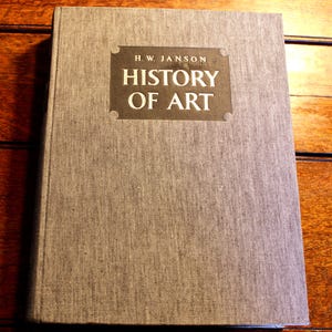 May include: A hardcover book titled "HISTORY OF ART" by H.W. Janson. The book has a gray linen cover with the title and author's name in white lettering on a dark brown rectangular label. The book is on a wooden surface.