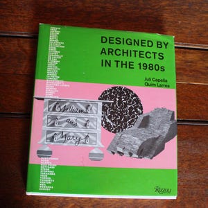 May include: Book cover with a green background and the title "Designed by Architects in the 1980s" by Juli Capella and Quim Larrea. The cover features a pink background with a black and white patterned chest of drawers, a black and white patterned circle, and a gray stone sculpture.