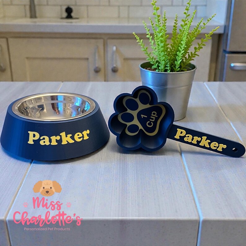 A navy blue dog bowl with a stainless steel insert and the name "Parker" in yellow letters is next to a navy blue paw-print shaped measuring cup with "1 Cup" and "Parker" on it.