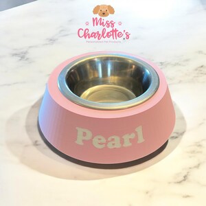 A light pink dog bowl with a stainless steel insert has the name "Pearl" in white letters on the front, sitting on a white marble surface.