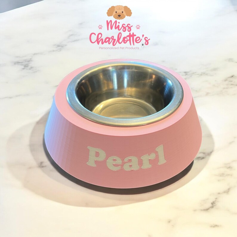 A light pink dog bowl with a stainless steel insert has the name "Pearl" in white letters on the front, sitting on a white marble surface.