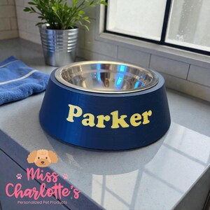 A navy blue dog bowl with a stainless steel insert has the name "Parker" in yellow letters on the front, sitting on a gray countertop.