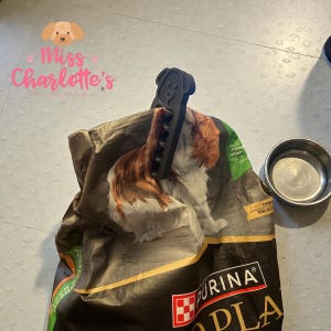 May include: A black dog-shaped bag clip secures a partially open bag of Purina dog food. The bag features a Cavalier King Charles Spaniel image. A stainless steel dog bowl sits nearby. The text "Miss Charlotte's" is visible.