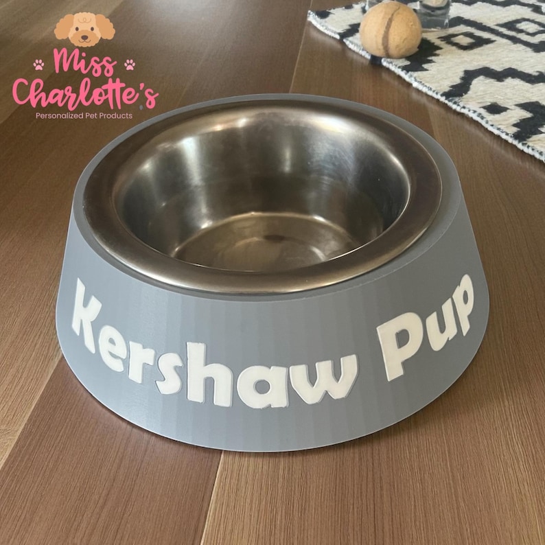 A light gray dog bowl with a stainless steel insert has the name "Kershaw Pup" in white letters on the front, sitting on a wooden floor next to a patterned rug.