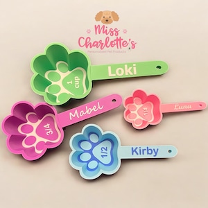 May include: A collection of five paw-shaped pet food measuring scoops in various colours: green, pink, purple, blue, and light pink. Each scoop is labelled with a pet's name and measurements in cups.