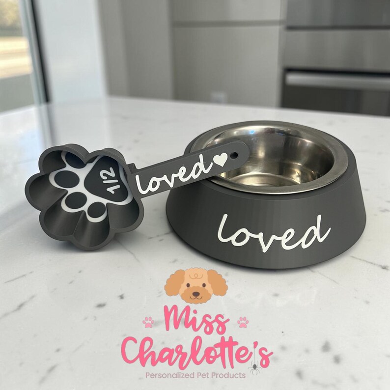 May include: A dark gray pet bowl with a matching paw-shaped measuring scoop. The bowl features the white text "loved" on the side. The scoop handle is labeled "loved" with a heart icon, and the scoop interior displays "1/2" in white text.