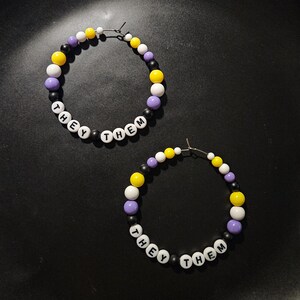 May include: A pair of hoop earrings made with purple, yellow, white, and black beads. The beads have the words "They Them" spelled out on them.