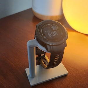 Op de afbeelding: Een zwart Garmin-horloge met een wit gezicht en een digitaal display dat de tijd 6:36 laat zien. Het horloge staat op een witte kunststof standaard.