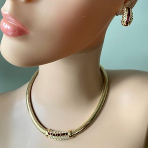 May include: A gold-tone metal necklace featuring a textured mesh chain and a central curved bar inlaid with a row of square-cut red stones flanked by clear crystals, paired with matching half-hoop earrings containing identical red and clear stones.