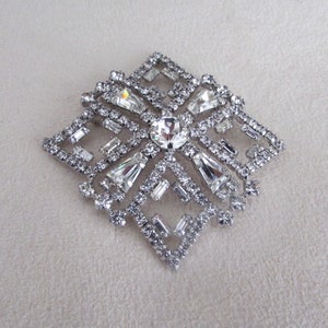 Peut inclure: Une broche argentée avec un motif géométrique orné de strass transparents. La broche est de forme carrée avec un motif en étoile au centre.