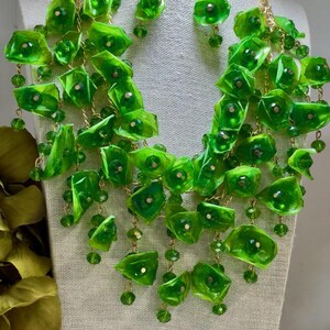 Puede incluir: Un collar de declaración verde y pendientes a juego con un diseño en cascada. El collar está hecho de flores de plástico verde y cuentas.