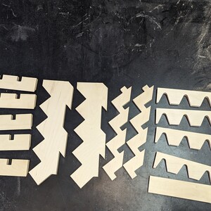 May include: A set of wooden pieces for a craft project. The pieces are cut in different shapes, including zigzags, waves, and rectangles. The pieces are made of light-colored wood and are arranged on a dark surface.
