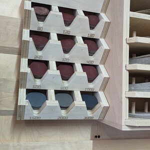 May include: A wooden wall-mounted organizer with slots for storing sanding discs. The slots are labeled with the grit numbers: 80, 120, 180, 240, 320, 400, 600, 900, 1000, 1500, 2000, and 3000.