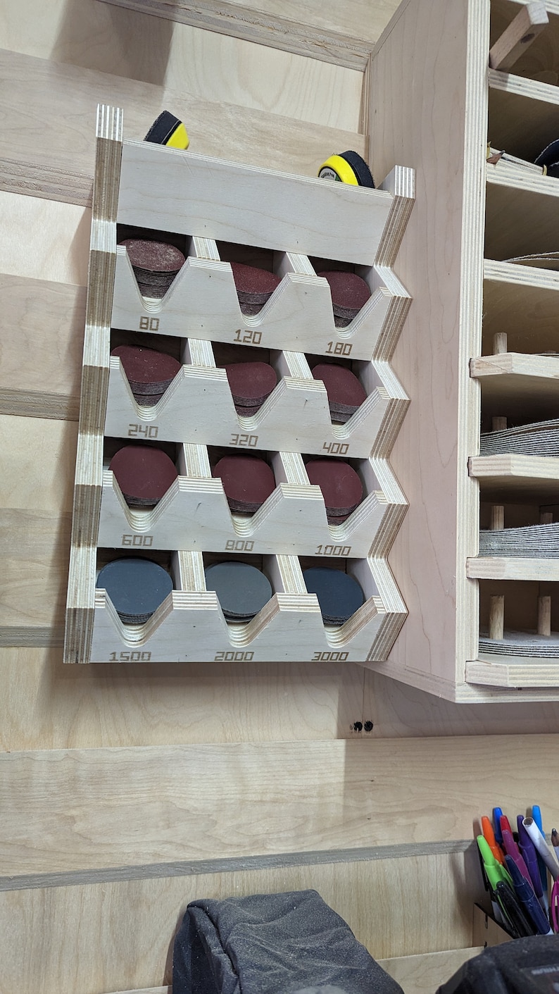 May include: A wooden wall-mounted organizer with slots for storing sanding discs. The slots are labeled with the grit numbers: 80, 120, 180, 240, 320, 400, 600, 900, 1000, 1500, 2000, and 3000.