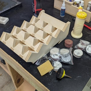 May include: A wooden sanding disc holder with numbered compartments for different grit sizes, from 80 to 3000 grit. There are also several bags of sanding discs in various sizes and colors.