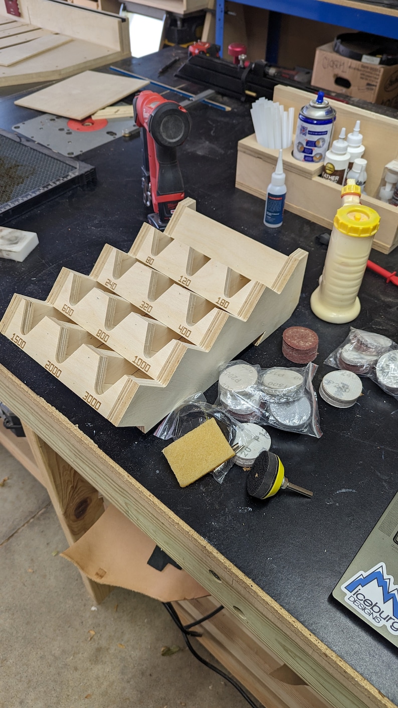 May include: A wooden sanding disc holder with numbered compartments for different grit sizes, from 80 to 3000 grit. There are also several bags of sanding discs in various sizes and colors.