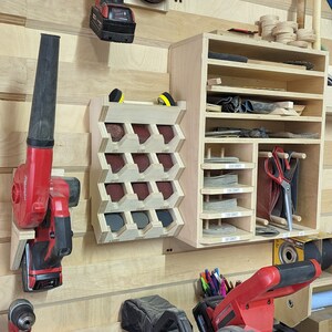 May include: A workshop wall with a variety of tools and storage solutions. A red and black power tool is mounted on the wall, along with a red and black leaf blower. A wooden storage shelf with multiple compartments holds various tools and supplies. A red and black power saw is on a table in the foreground.