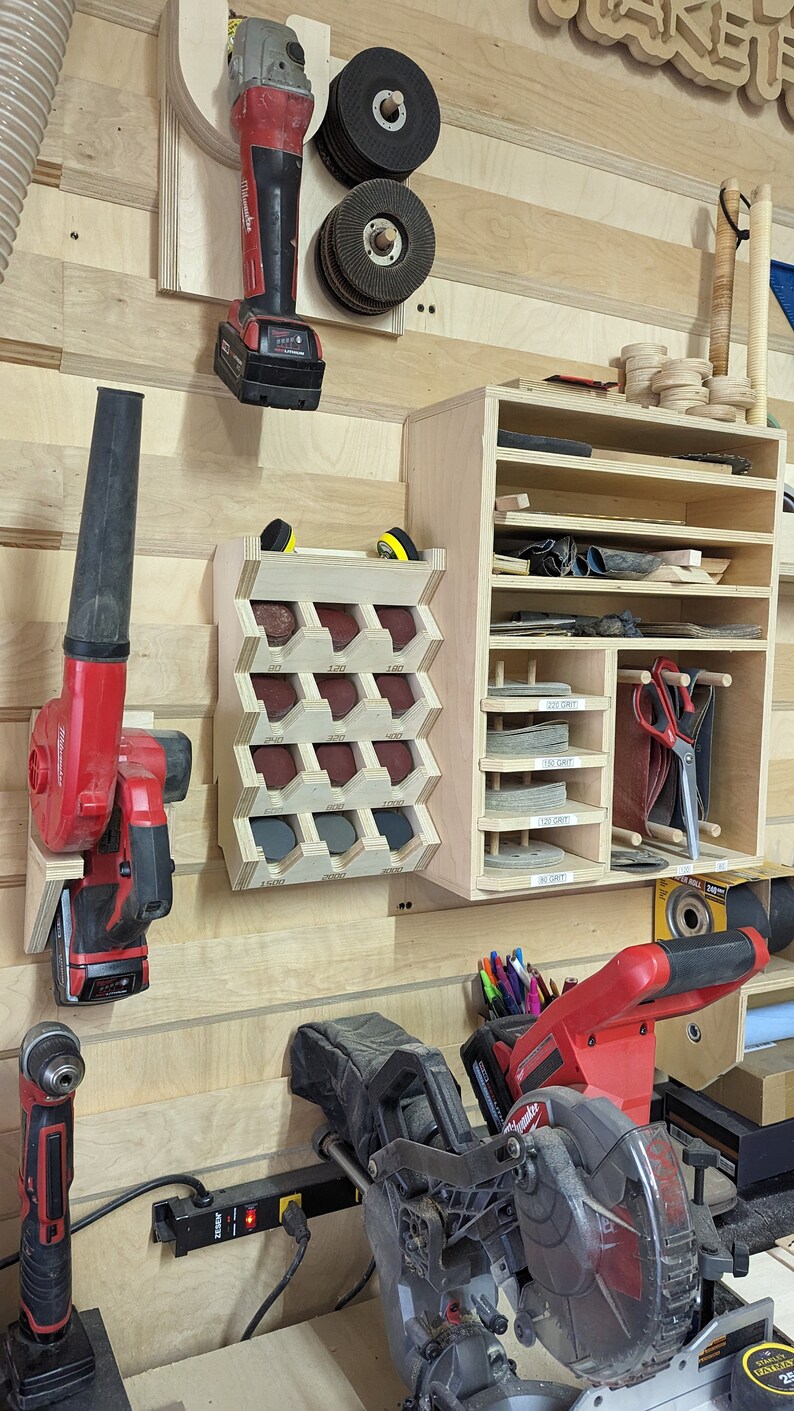 May include: A workshop wall with a variety of tools and storage solutions. A red and black power tool is mounted on the wall, along with a red and black leaf blower. A wooden storage shelf with multiple compartments holds various tools and supplies. A red and black power saw is on a table in the foreground.