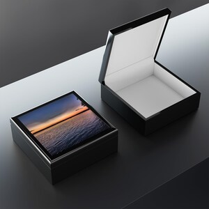 May include: Two black rectangular jewellery boxes, one open and one closed. The closed box has a lid with a scenic image of a sunset over water. The open box reveals a white interior. Both boxes have a glossy finish.
