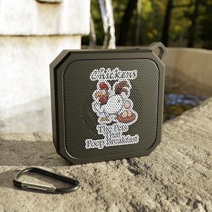 May include: A black, square portable speaker with a carabiner clip. The speaker has a graphic of a cartoon rooster and the text "Chicken's The Best Poop Breakfast". The speaker is sitting on a stone surface.