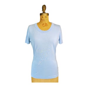 Puede incluir: Una camiseta de manga corta en color azul claro con cuello redondo y dobladillo recto. La prenda se muestra sobre un maniquí de costura de color beige. El tejido parece ser un material de punto fino con una textura suave y uniforme.