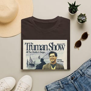 Könnte beinhalten: Ein braunes T-Shirt mit einem Grafikdesign aus dem Film "The Truman Show". Die Grafik enthält den Filmtitel, den Satz "All The Worlds A Stage" und ein Bild von Jim Carrey. Das Shirt wird mit einem Strohhut, einer Sonnenbrille und Jeans kombiniert.