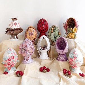 Puede incluir: Una colección de huevos de Pascua decorativos de cerámica en varios colores y diseños. Algunos huevos presentan motivos florales, mientras que otros tienen calados o motivos de mariposas. Los huevos se exhiben sobre una tela color crema.