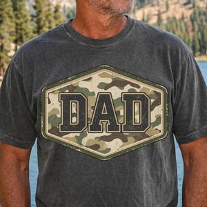 May include: Dark gray t-shirt with a camouflage "DAD" graphic. The word "DAD" is in large, block letters, set against a camouflage pattern within a hexagonal frame. The shirt is short-sleeved and appears to be made of a soft, worn material.