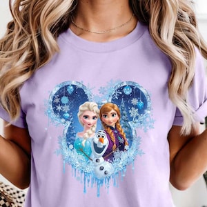 May include: A light purple t-shirt featuring a graphic of Elsa, Anna, and Olaf from the movie Frozen. The design is set within a Mickey Mouse-shaped frame with snowflakes and icicles.