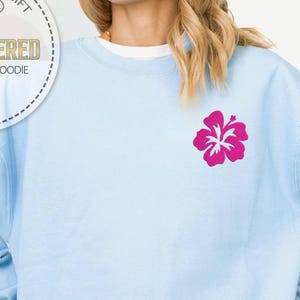 Puede incluir: Sudadera azul claro con una flor de hibisco fucsia bordada en el pecho. La sudadera tiene mangas largas y cuello redondo. La imagen tambi&eacute;n muestra un logotipo circular que dice "Regalo Personalizado" y "Camiseta y Sudadera con Bordado".