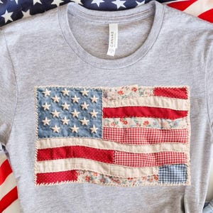 May include: A heather grey short-sleeve t-shirt featuring a rectangular patch of the American flag. The flag design is constructed from various fabric scraps in red, white, and blue, including floral and gingham patterns, with embroidered stars.