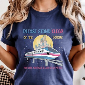 May include: Navy blue t-shirt featuring a monorail design with a castle and the text "PLEASE STAND CLEAR OF THE DOORS" in colorful letters. The design includes the text "POR FAVOR, MANTENGASE ALEJADO DE LAS PUERT" at the bottom.