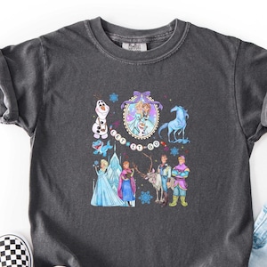 May include: Dark gray t-shirt featuring a colorful graphic of characters from the movie "Frozen", including Elsa, Anna, Olaf, and Kristoff. The phrase "Let It Go" is also displayed. The shirt is paired with black and white checkered slip-on shoes.