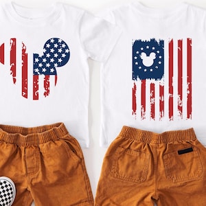 May include: Two white t-shirts with red, white, and blue American flag designs, one in the shape of a mouse head and the other a flag. Paired with orange shorts and black and white checkered slip-on shoes. A patriotic children's outfit.